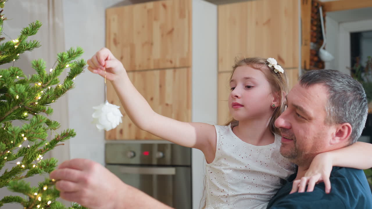Close up view of caring dad carrying daughter in arms while she carefully hangs white ornament on christmas tree inside cozy festive decorated kitchen during cheerful family holiday celebration