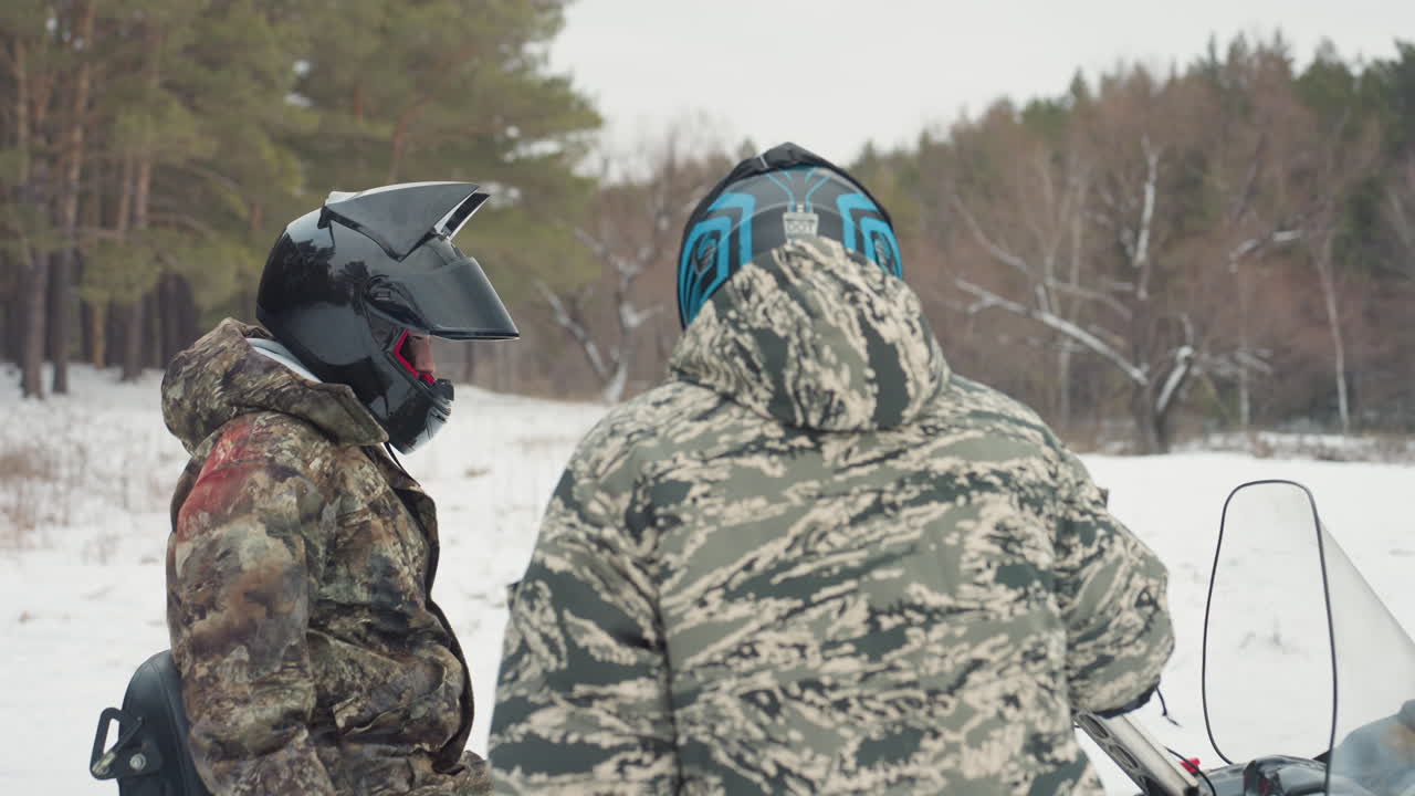 Two riders camo helmets checking atv in snowy clearing, team leader inspects throttle while partner adjusts gloves, mechanic role inspects engine, utility vehicle parked in background, focused