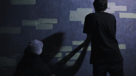 two males work on a mural painting green bricks onto a purple wall to form graffiti