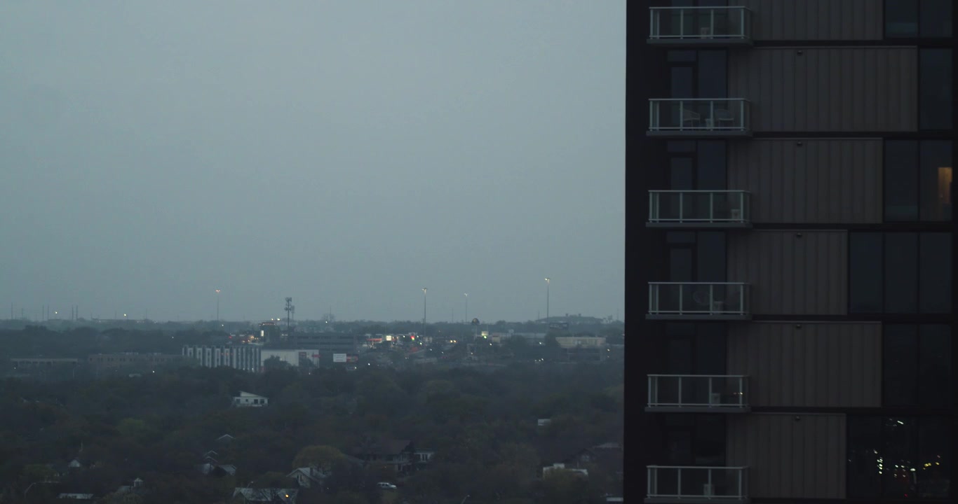 Vista elevada e nebulosa de Austin, Texas, ao entardecer, mostrando um arranha-céu residencial contra um horizonte urbano distante e luzes da cidade suaves.