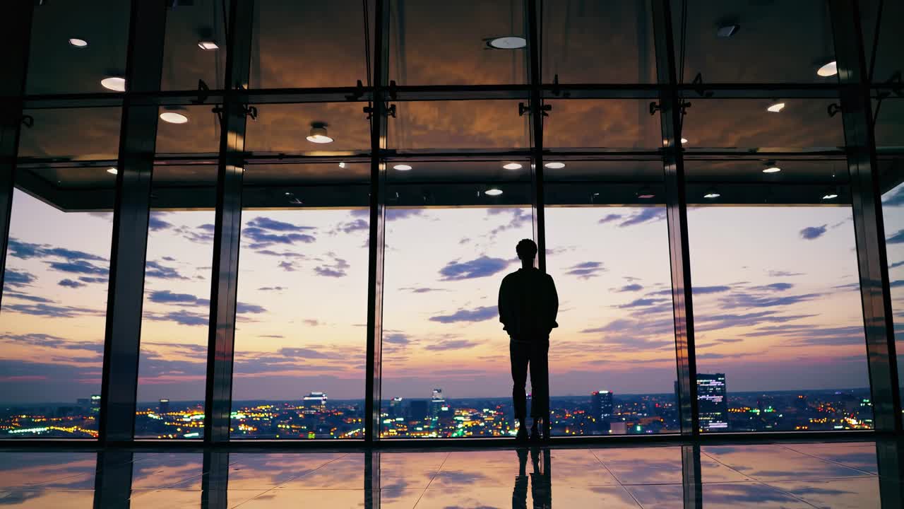 Silhouette of a person standing by large windows, overlooking a cityscape at sunset