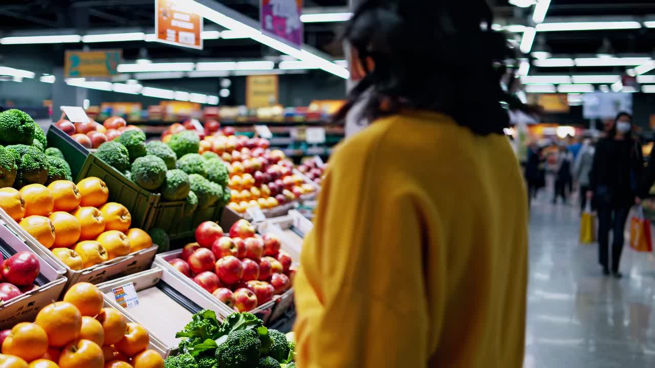 A woman in a yellow sweater selects apples in a supermarket. The video captures a side angle