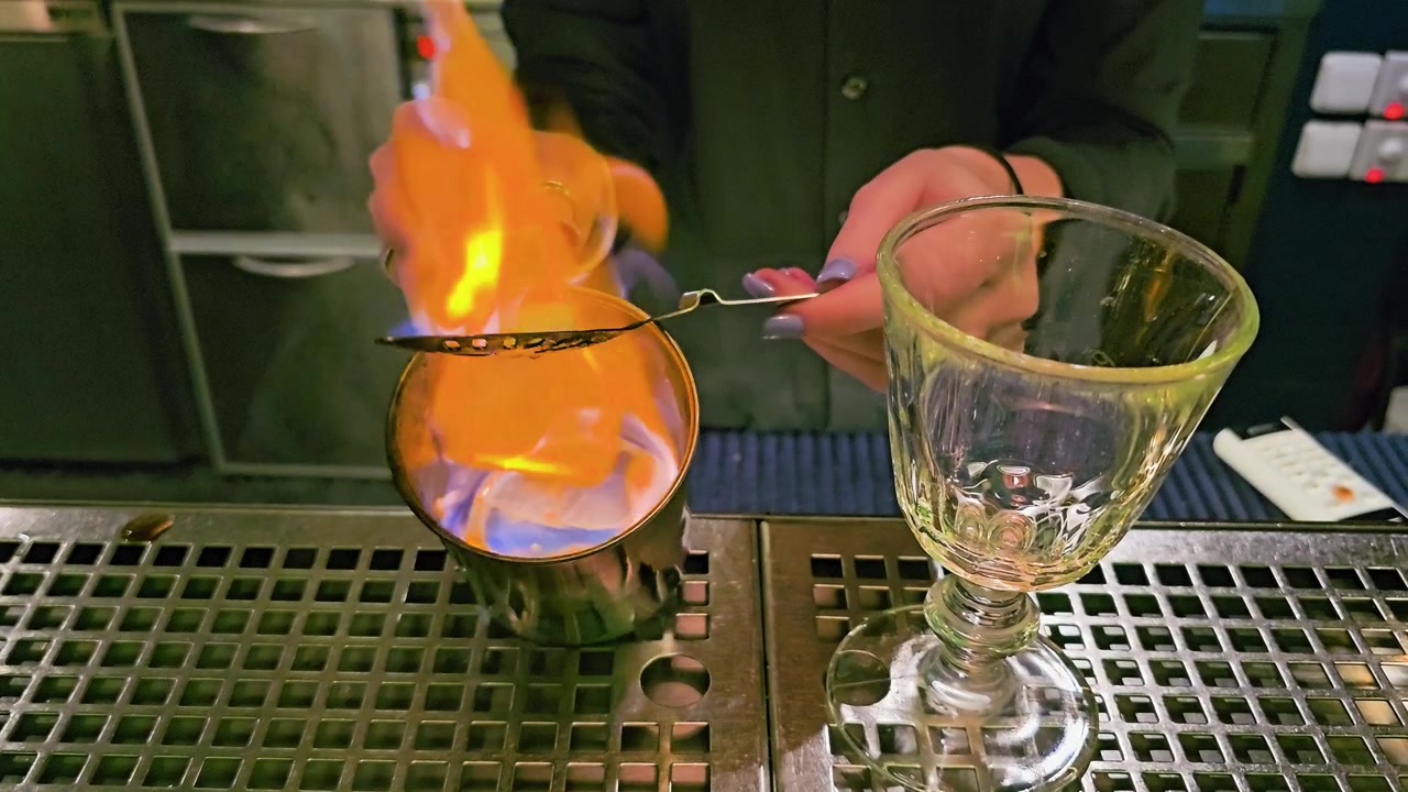 Scena di mixology suggestiva con un cubetto di zucchero all'assenzio fiammeggiante, cucchiaio perforato, tazza di metallo, calice vuoto, vassoio raccogligocce in acciaio inossidabile, mani del barista e illuminazione scura del bar di notte.