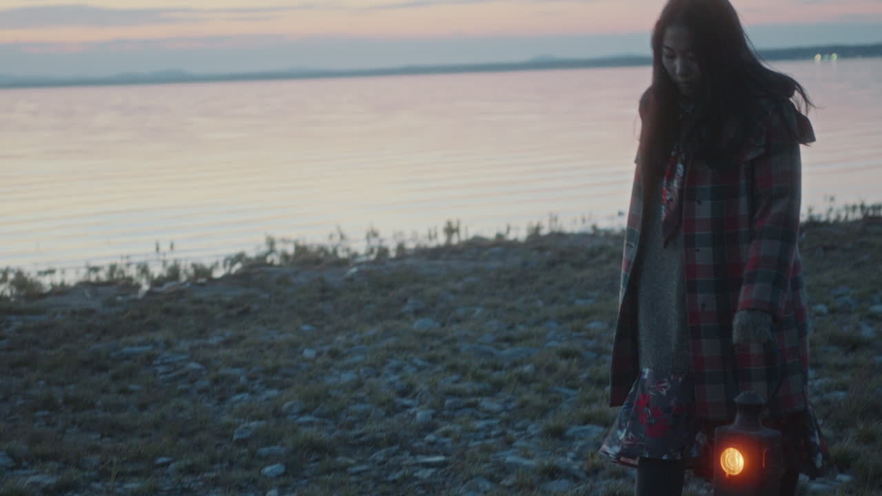 Asian Girl with Flickering Lantern Walking along Rocky Lakeshore at Dusk