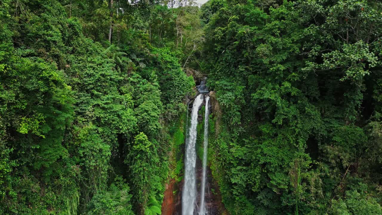 Drone footage of Gitgit Waterfall in Bali, captured from a closer angle, showcasing powerful tropical water flow surrounded by dense green trees and foliage in a vibrant natural setting