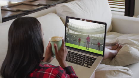compuesto de una mujer sentada en casa sosteniendo un café viendo un partido de rugby en una computadora portátil