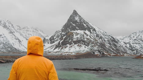 man in yellow hooded jacket admiring famous bridge and beautiful snowy landscape of the lofoten islands - wide shot