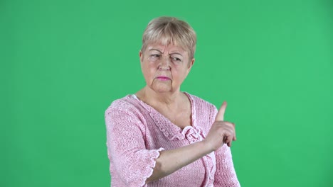 카메라를 바라보는 노인이 누군가에게 비난하고 지표손가락을 흔들며 위협합니다. 스튜디오에서 녹색 스크린에서 분홍색 스웨터를 입은 짧은 머리카락을 가진 회색 머리카락의 할머니.