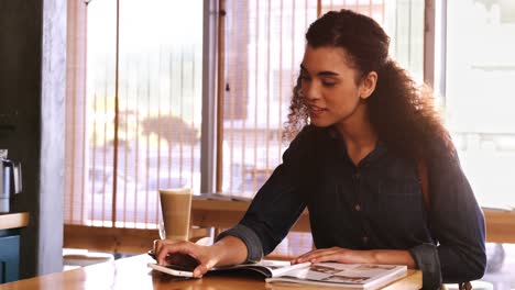 Mujer-Usando-Teléfono-Móvil-Mientras-Lee-Una-Revista-En-Una-Cafetería