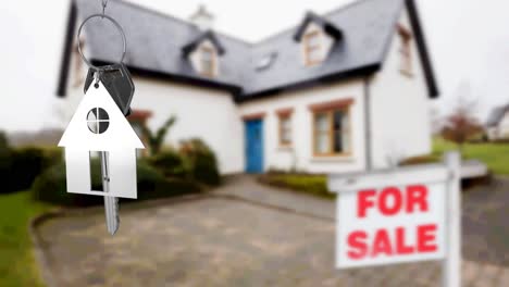 Animación-Del-Llavero-Y-La-Llave-Plateados-De-Una-Casa,-Colgados-Frente-A-Una-Casa-Borrosa-Con-Un-Cartel-De-Venta