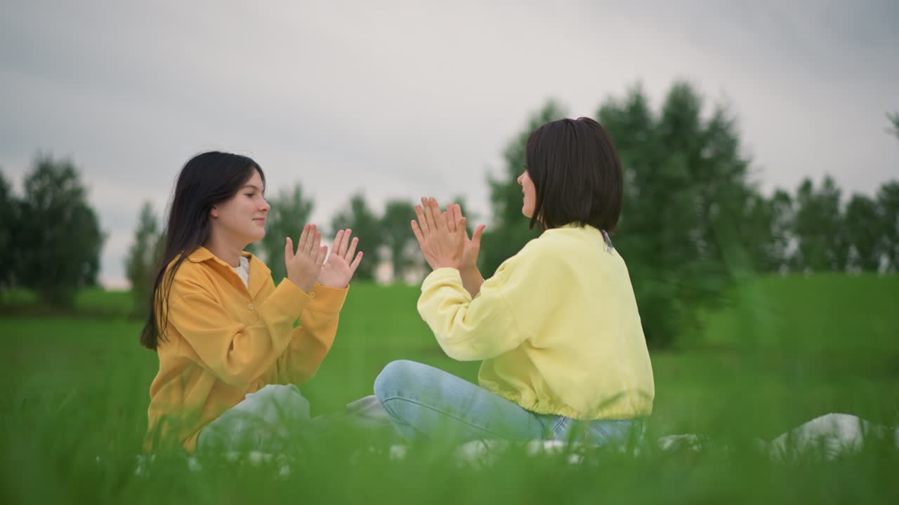 緑の牧草地に座り、黄色いジャケットとデニムを着て、互いに向き合って草の上に座り、手をたたいて笑っている2人の女性、柔らかな曇り空、遠くの木々、親密で遊び心のある雰囲気、子供時代の思い出