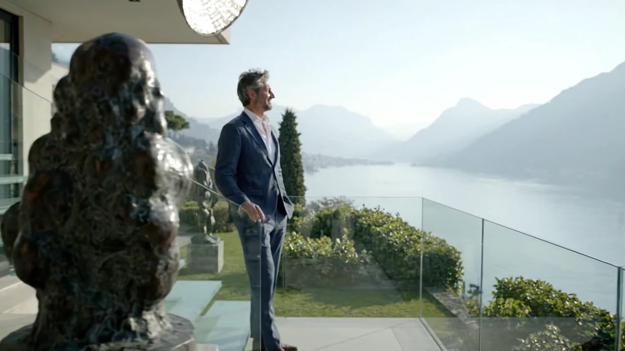 Un uomo elegante in un abito impeccabile si gode la bellezza panoramica di una vista sul lungomare da una moderna scala in vetro, che si affaccia su montagne e acqua serene.