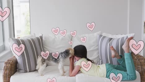 animation of hearts moving over caucasian girl wearing face mask playing with her pet dog