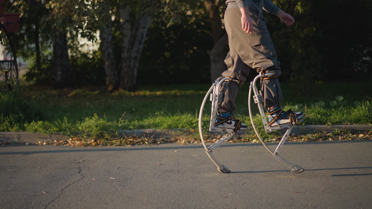 Guy walking with stilts wearing gray hoodie and pants on paved road surrounded by green grass and trees, showcasing dynamic movement, balance, and outdoor activity, emphasizing agility