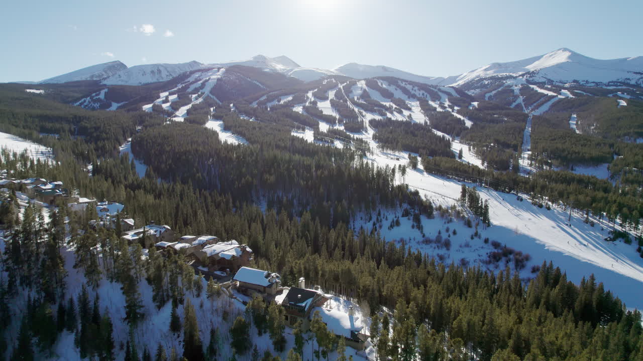 An epic aerial reveal of the popular Breckenridge ski resort in Colorado, with snow covered mountain peaks and high altitude trails full of winter skiers and snowboarders on a sunny winter day.