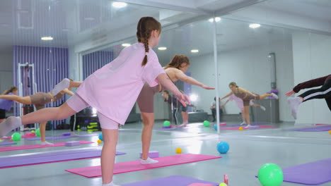 group training session in bright fitness studio with participants balancing on one leg with arms open, colorful exercise balls scattered around, large mirror reflecting their movements