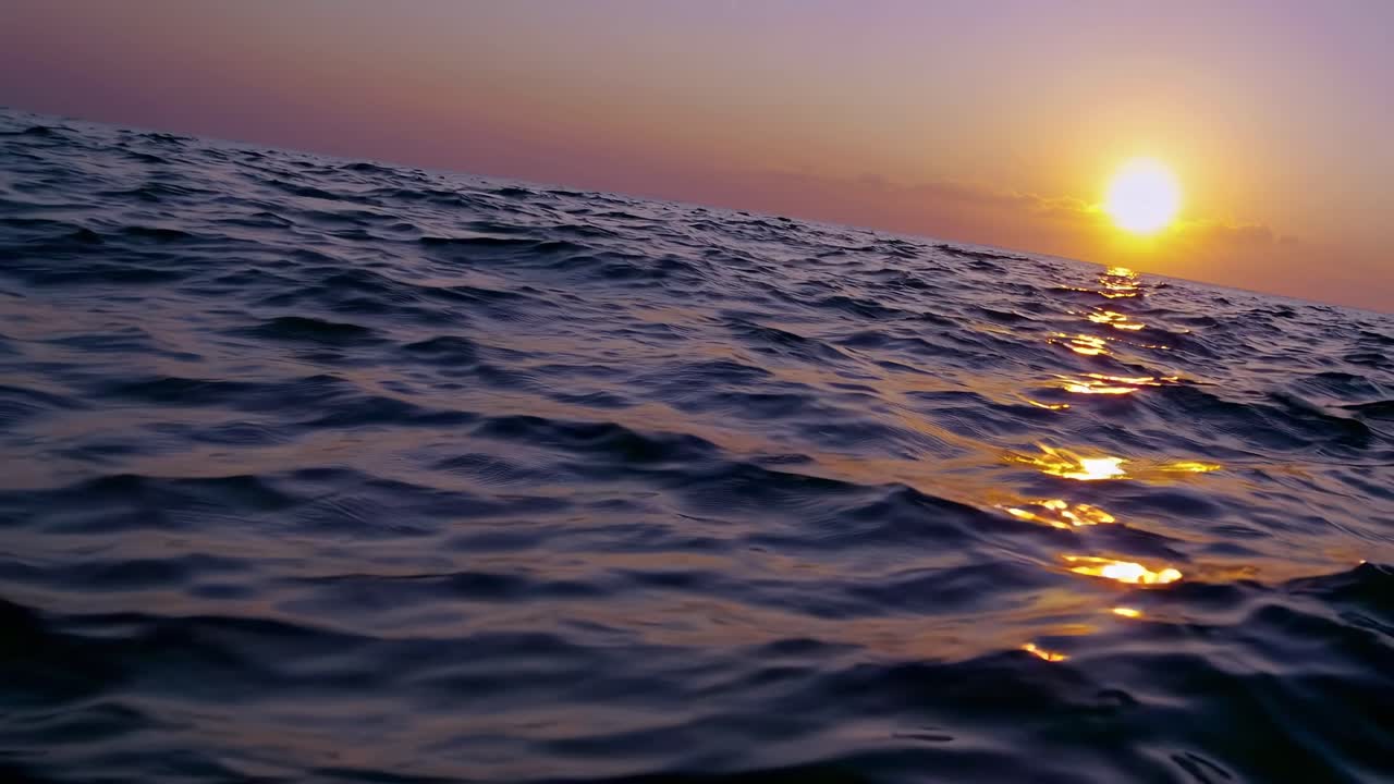A tilted low-angle video shot captures the serene ocean waves at sunset, with warm hues reflecting
