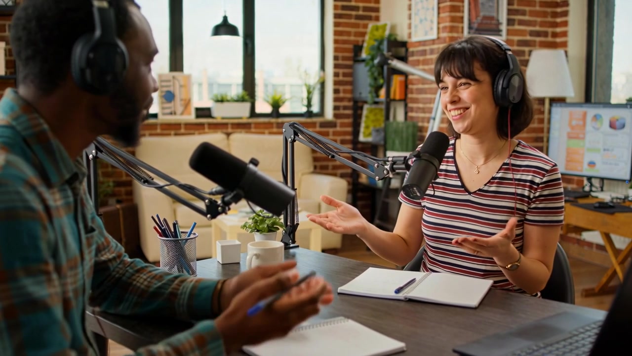 Conduttori che registrano, un uomo che prende appunti e parla in un microfono a giraffa mentre una donna gesticola in uno studio di podcast.