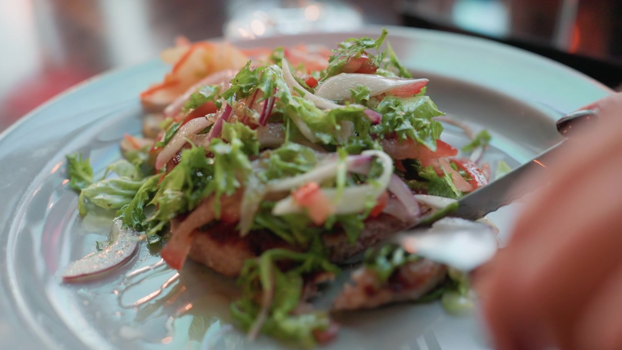 Hand cutting into fresh salad greens and grilled meat on white plate with visible ingredients like lettuce tomatoes onions. Soft lighting highlights texture details in indoor dining setting