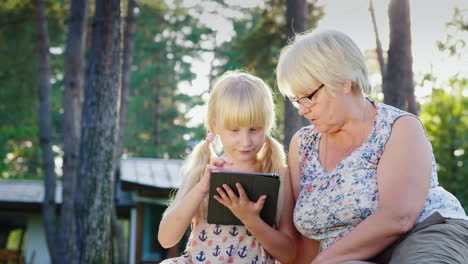 Großmutter-Und-Enkelin-Ruhen-Sich-Im-Hinterhof-Des-Hauses-Aus,-Sie-Schauen-Gemeinsam-Auf-Das-Tablet