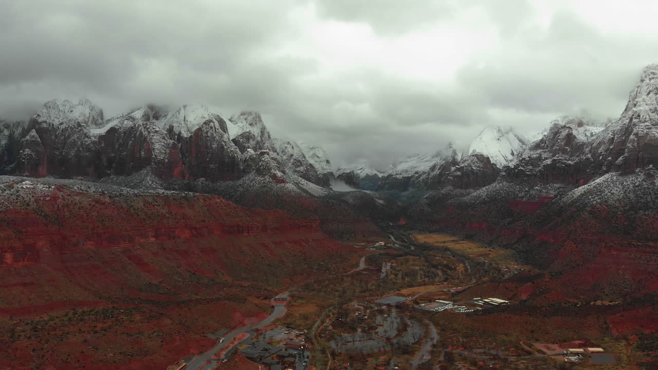 Aerial shot of the red rock mountains in Utah with snow on the peaks sheltering a small village in between them on a cloudy day.