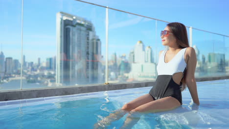 sexy young asian girl sitting on swimming pool edge moves legs in water looking at cityscape