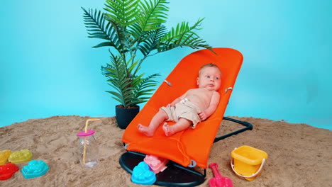 beautiful baby boy wearing shorts lies in orange chair. kid sits on the sand with toys and plant in pot beside.