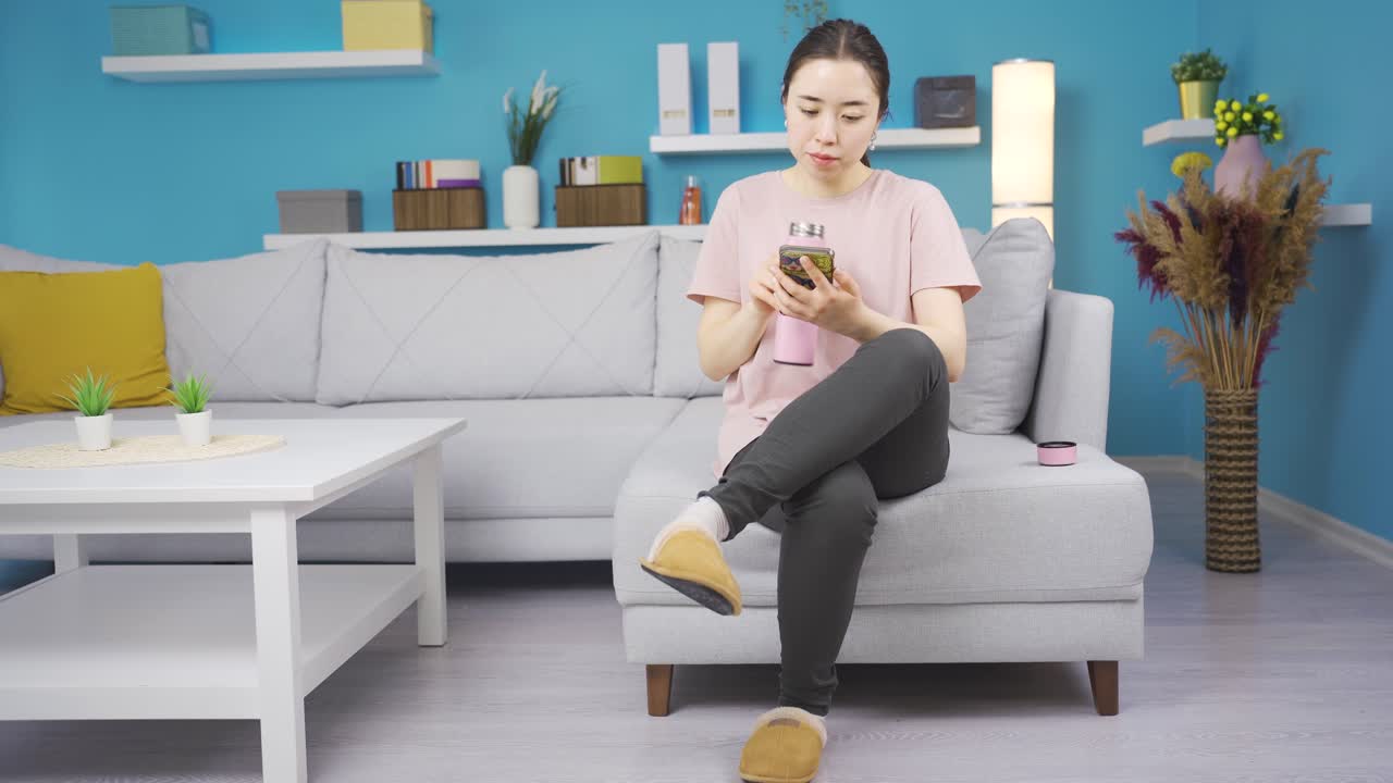 슬리퍼를 입은 아름다운 아시아 여성이 집에서 테르모스에서 음료를 마시고 있습니다.