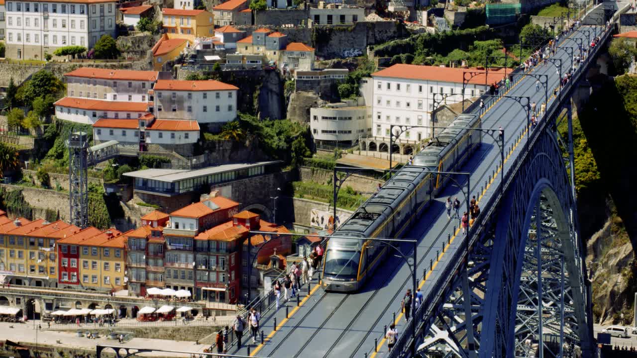 Turisti e metropolitana che attraversano il Ponte Dom Luís I con folle e bancarelle del mercato sottostanti, Porto, Portogallo. Ripresa aerea cinematografica in 4K al rallentatore di una città mediterranea estiva.