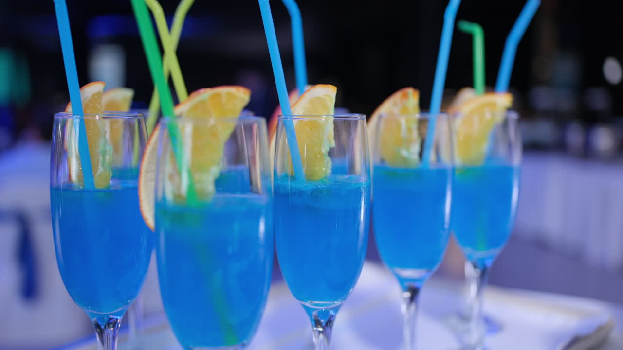 Multiple glasses with a blue cocktail, garnished with orange slices and colorful straws, on a tray