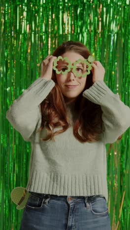 vertical video of woman celebrating st patrick's day standing in front of green tinsel curtain wearing prop shamrock shaped glasses