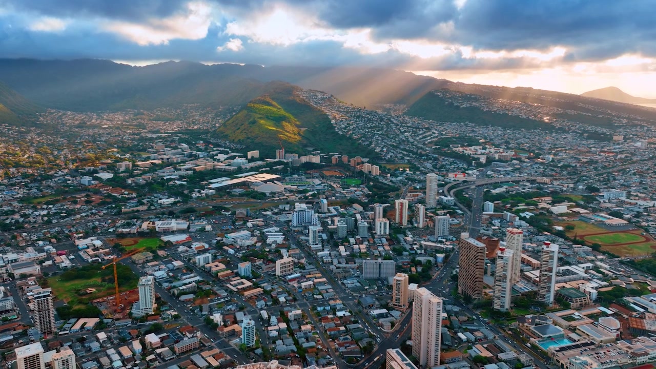 I raggi del sole al tramonto attraversano le nuvole grigie illuminando le montagne. Volo con drone sopra il denso paesaggio urbano di Honolulu, Hawaii, USA