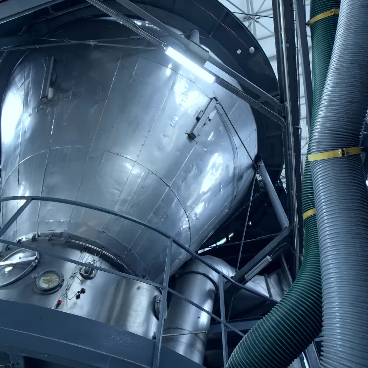 Huge dairy tank beside the thick plastic tubes. Stainless steel tank standing at the raised platform with sensors at its basics. Circle view from below