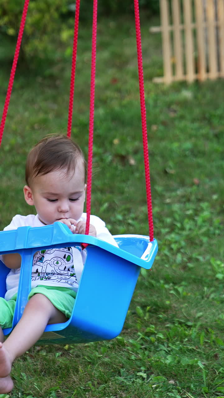 Little boy swaying in a swing looking attentively at the fastener in his hands. Cute kid in the summer garden. Nature backdrop. Vertical video