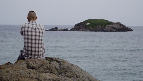 Caucasian-millennial-sit-alone-on-rock-by-island-sea-shore,-gloomy-overcast-day