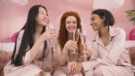 front view of three multiethnic friends toasting with champagne sitting on sofa celebrating bachelorette party 1