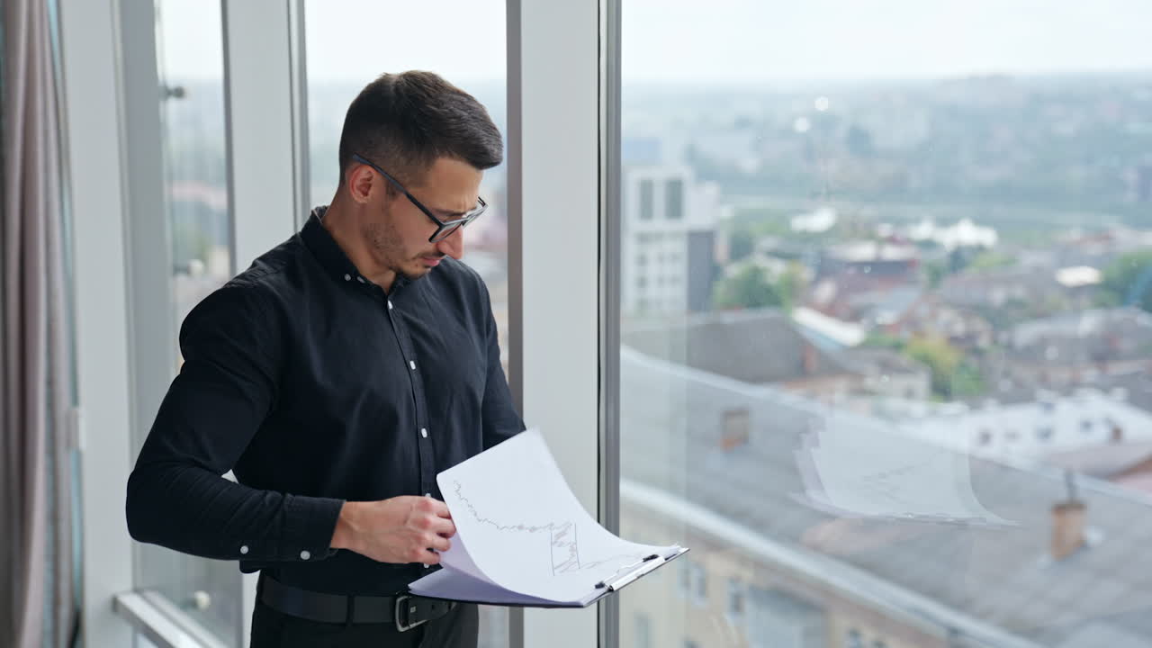 Concerned Caucasian man looking through the papers standing at the window. Cityscape in windows at backdrop in blur.
