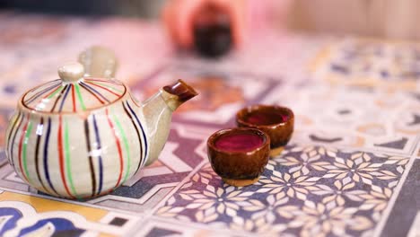 a hand pours tea from a striped teapot into small cups on a patterned tablecloth, creating a warm, inviting atmosphere