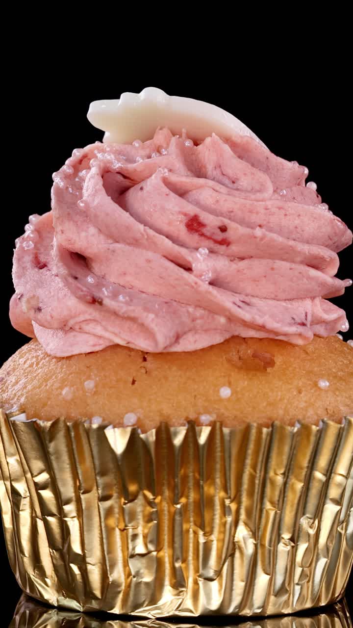 Un cupcake a tema fragola con glassa rosa e un topper decorativo ruota fluidamente su uno sfondo nero, illuminato da un'illuminazione da studio brillante e uniforme.