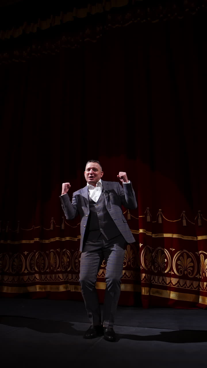 Actor in suit on large theater stage. Male comedian telling jokes and dancing gestures on the scene. Joyful performer on stage. Vertical video