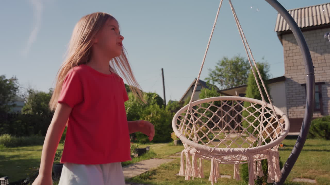 Energetic Girl Dances Near Swinging Chair Outside, Young Girl Reaches Up Playfully By Outdoor Swing In Sunlight, Child Enjoying Childhood Moments By Sunny Backyard With Swing And Green Surroundings