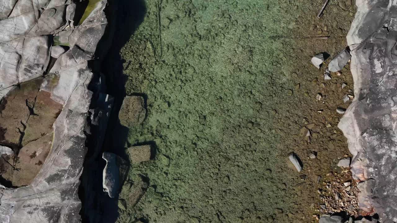 Vertical drone footage studies a quiet rockpool set into black shoreline rocks, its clear, calm water gently reflecting the sky as small ripples form around the rugged coastal edge
