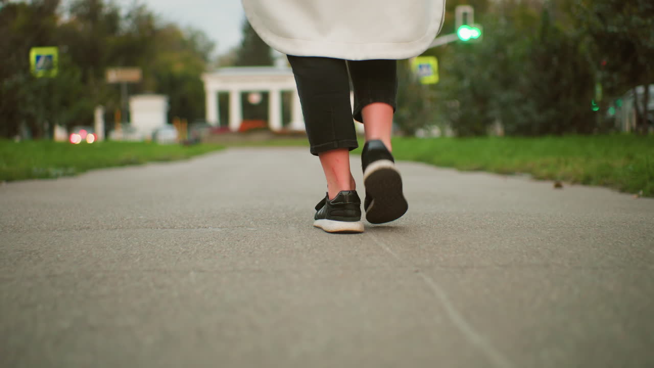 Leg view of person wearing long coat and black sneakers walking along urban sidewalk while twirling closed umbrella, another person passing by, blurred greenery and city structures in soft background