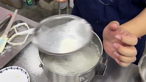 Top-down-view,-pastry-chef-gently-patting-on-the-side-of-strainer,-fine-and-fluffy-cake-flour-and-caster-sugar-sieving-through-into-the-stainless-steel-mixing-bowl