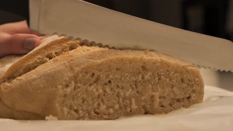 Mano-Femenina-Sosteniendo-Y-Cortando-Pan-De-Masa-Agria-Recién-Horneado-Con-Un-Cuchillo-De-Cocina-Afilado-En-La-Mesa