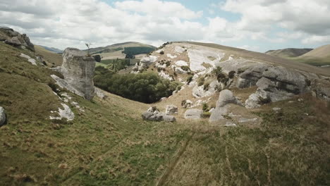 Forward-drone-shot-of-rocky-hills-covered-in-grass,-in-New-Zealand,-with-some-meadows-and-trees,-on-a-cloudy-day