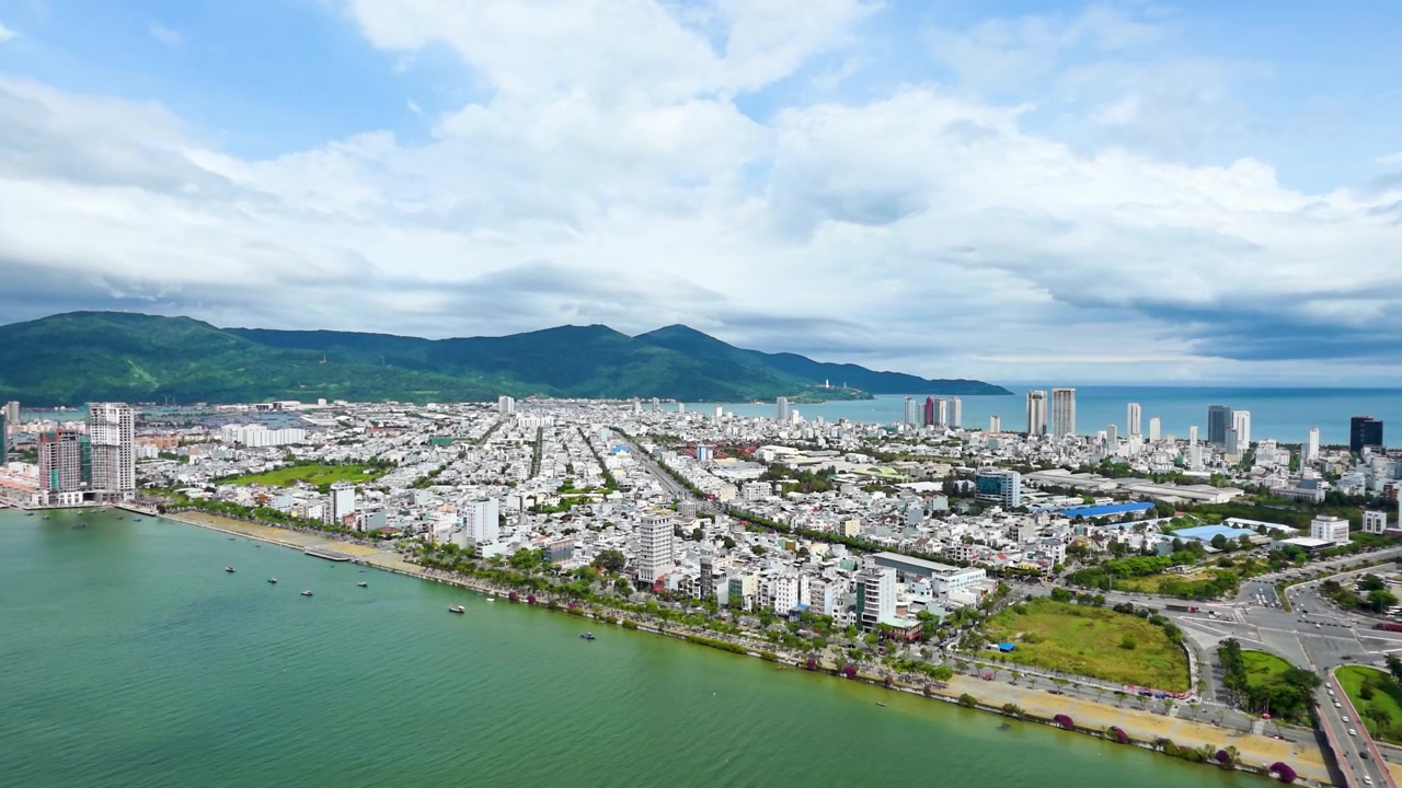 Cảnh quay trên không đầy nắng, phóng to qua thành phố Đà Nẵng và sông Hàn xanh ngọc bích hướng về bán đảo Sơn Trà xanh tươi với Biển Đông lấp lánh ở phía sau