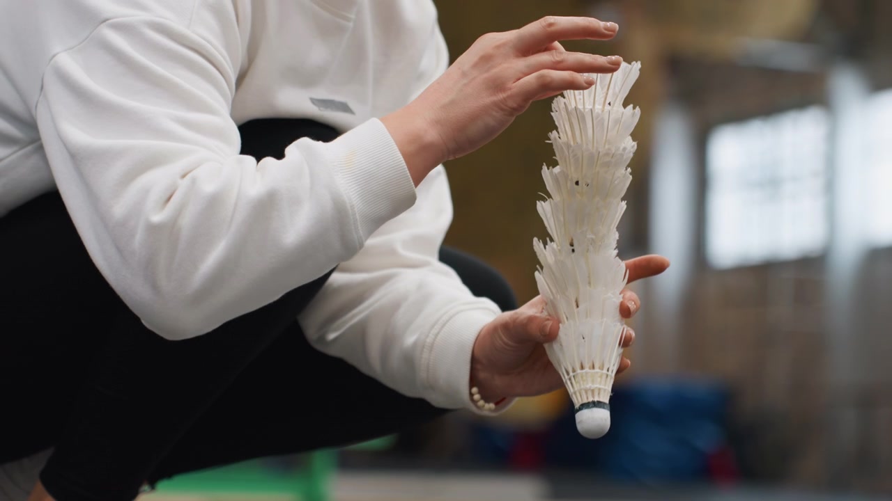 Player collecting shuttlecocks on court, hands stacking stacks of feathered shuttles and arranging pile, indoor sports hall scene with coach nearby, quick warmup and drill setup, communal practice