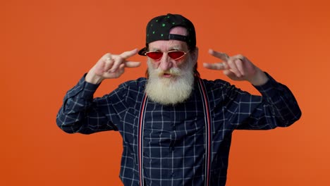 un hombre mayor con barba blanca, gafas de sol y un sombrero baila con un signo de paz