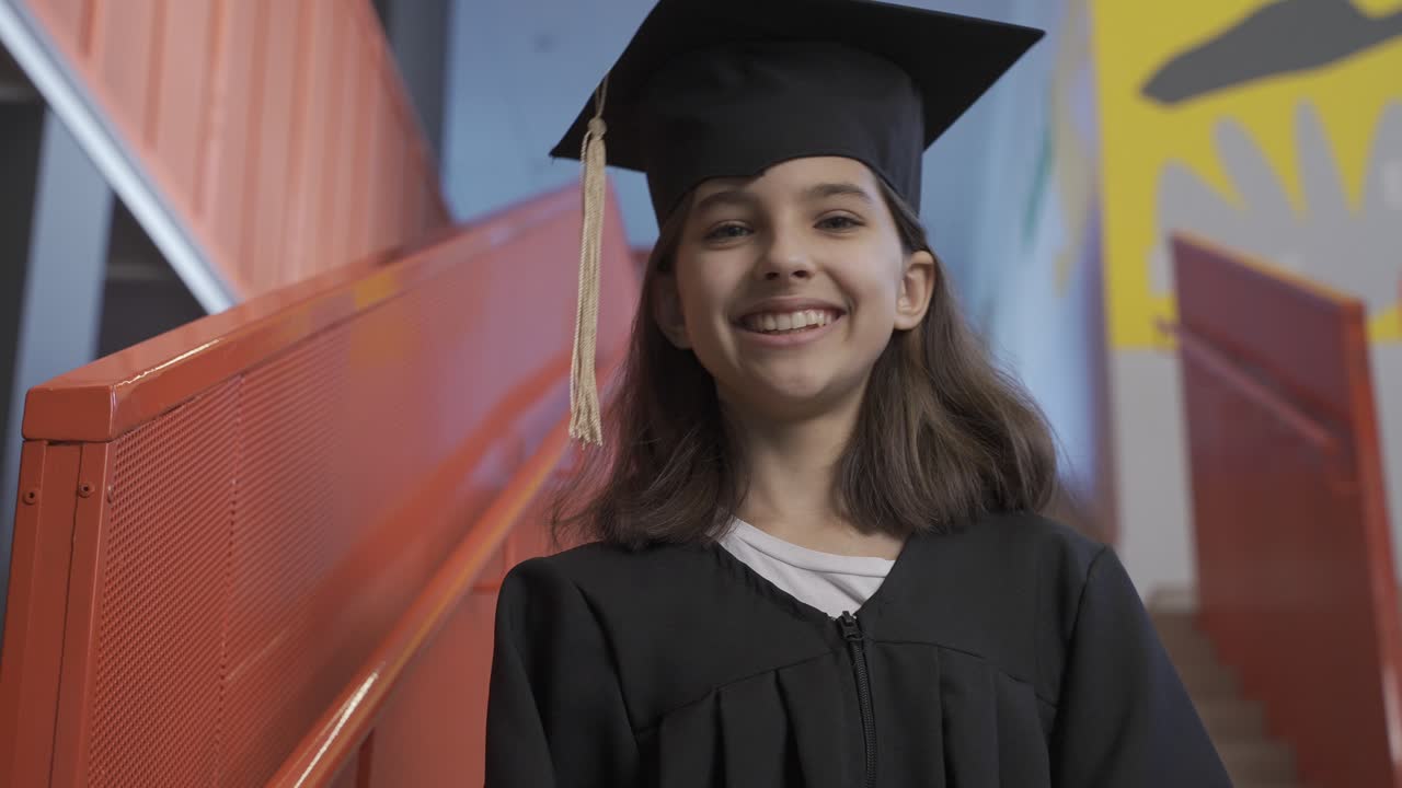 retrato de una feliz estudiante de preescolar con toga y birrete sosteniendo un diploma de graduación y mirando la cámara